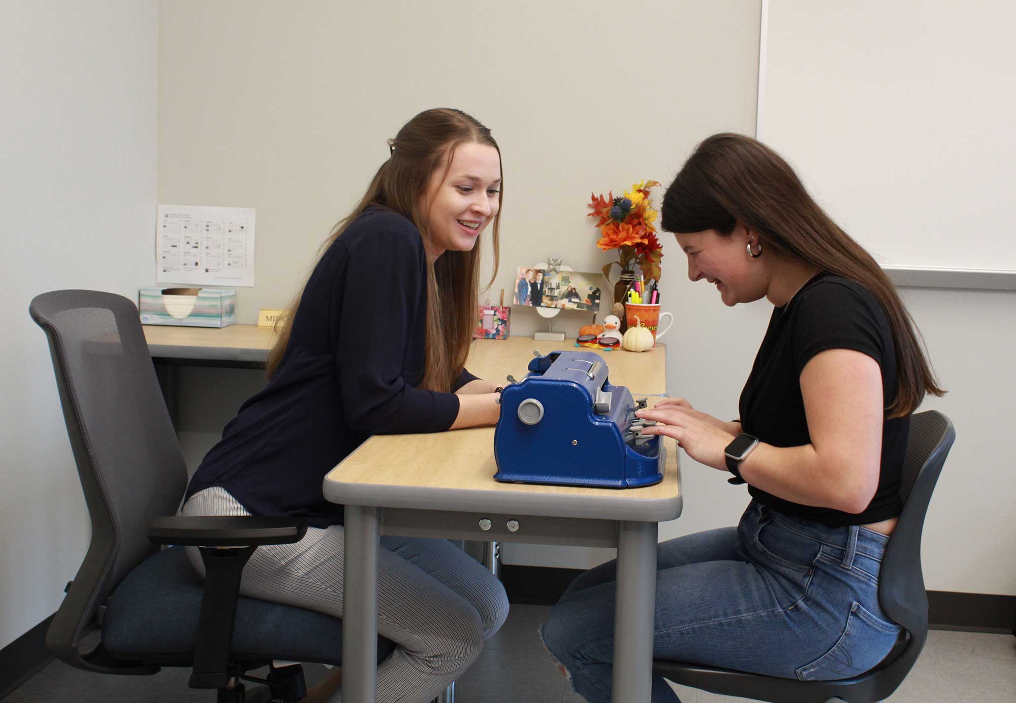 Two young women sit across from each other at a table in a classroom. One, wearing a navy blue long-sleeve shirt, smiles while watching the other, who wears a black T-shirt and jeans, type on a blue Braille writer. The classroom features a whiteboard, a desk, and fall-themed decorations on a shelf in the background.