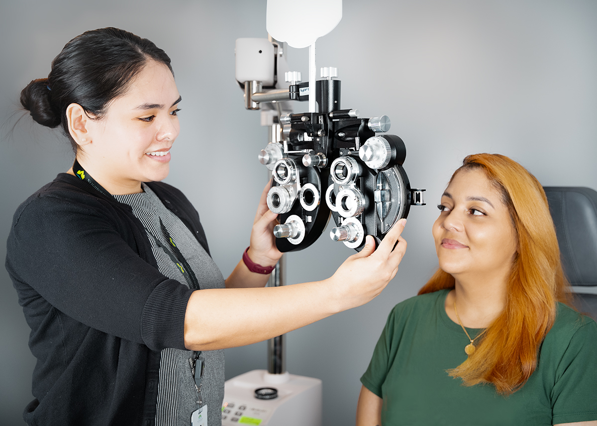 A woman in a green shirt undergoing a vision screening. The optometrist uses a large phoropter device to determine the woman's prescription.