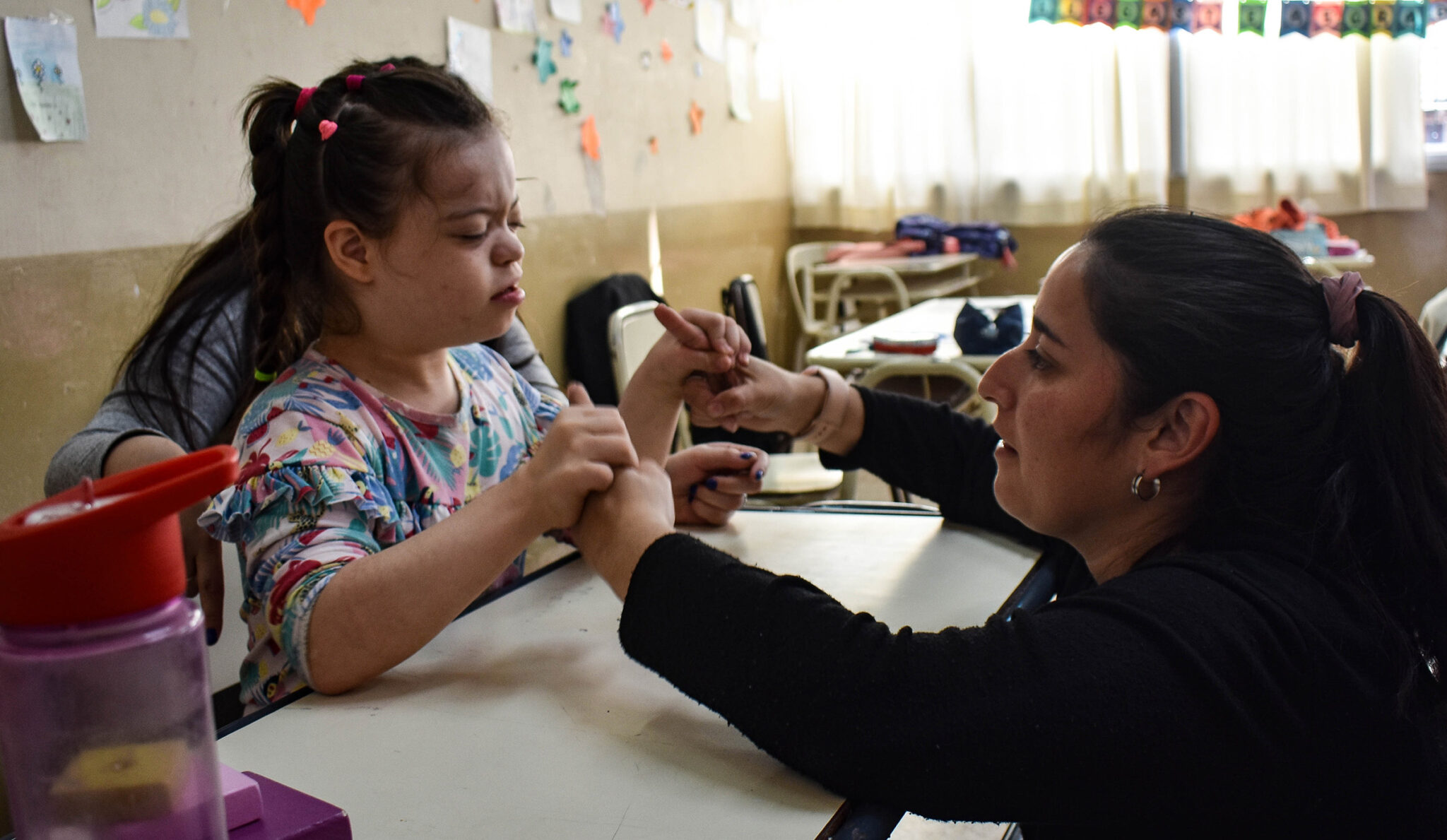 Vicky, a sociable student who is deafblind and with multiple disabilities, is using tactile Sign Language to communicate with her teacher. In the image, Vicky places her hands gently on top of the teacher’s, allowing her to feel the shape and movement of the teacher's hands. This tactile exchange helps Vicky understand that they are about to head to music class. The background of the classroom includes desks, school supplies, and decorations, creating a typical learning environment.