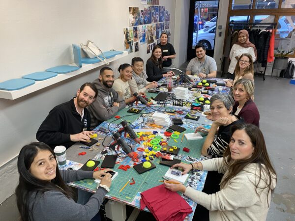 A diverse group of 13 people are positioned around a large table filled with colorful plastic shapes, cutting mats, glue guns, and craft materials in the process of creating communications cards.