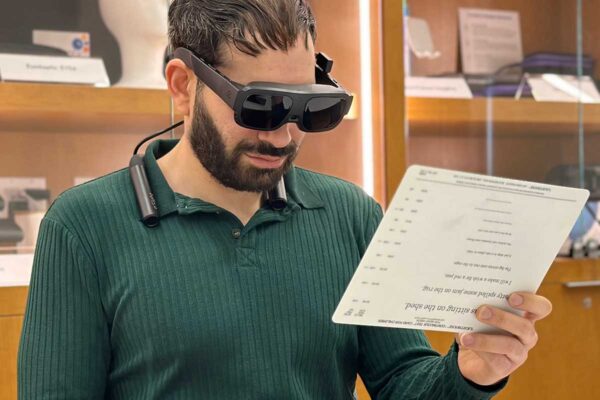 A man in a green shirt is wearing an assistive technology device that looks like a pair of goggles. He is holding up a sheet of paper with text on it.
