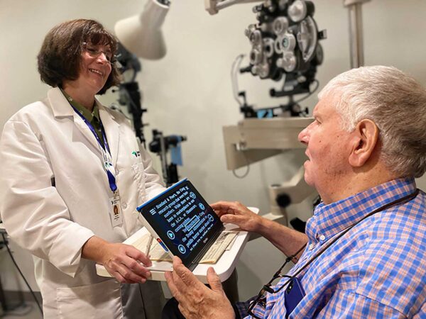 A man is holding a portable electronic magnifier with text written on the screen. A doctor in a white lab coat is standing next to him, smiling.