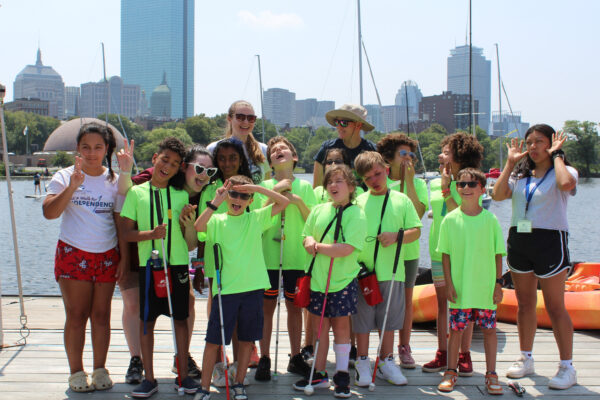 A group of children in bright neon green shirts and counselors stand on a dock with the Boston city skyline in the background. They are holding white canes and some children are making peace signs or playful gestures.